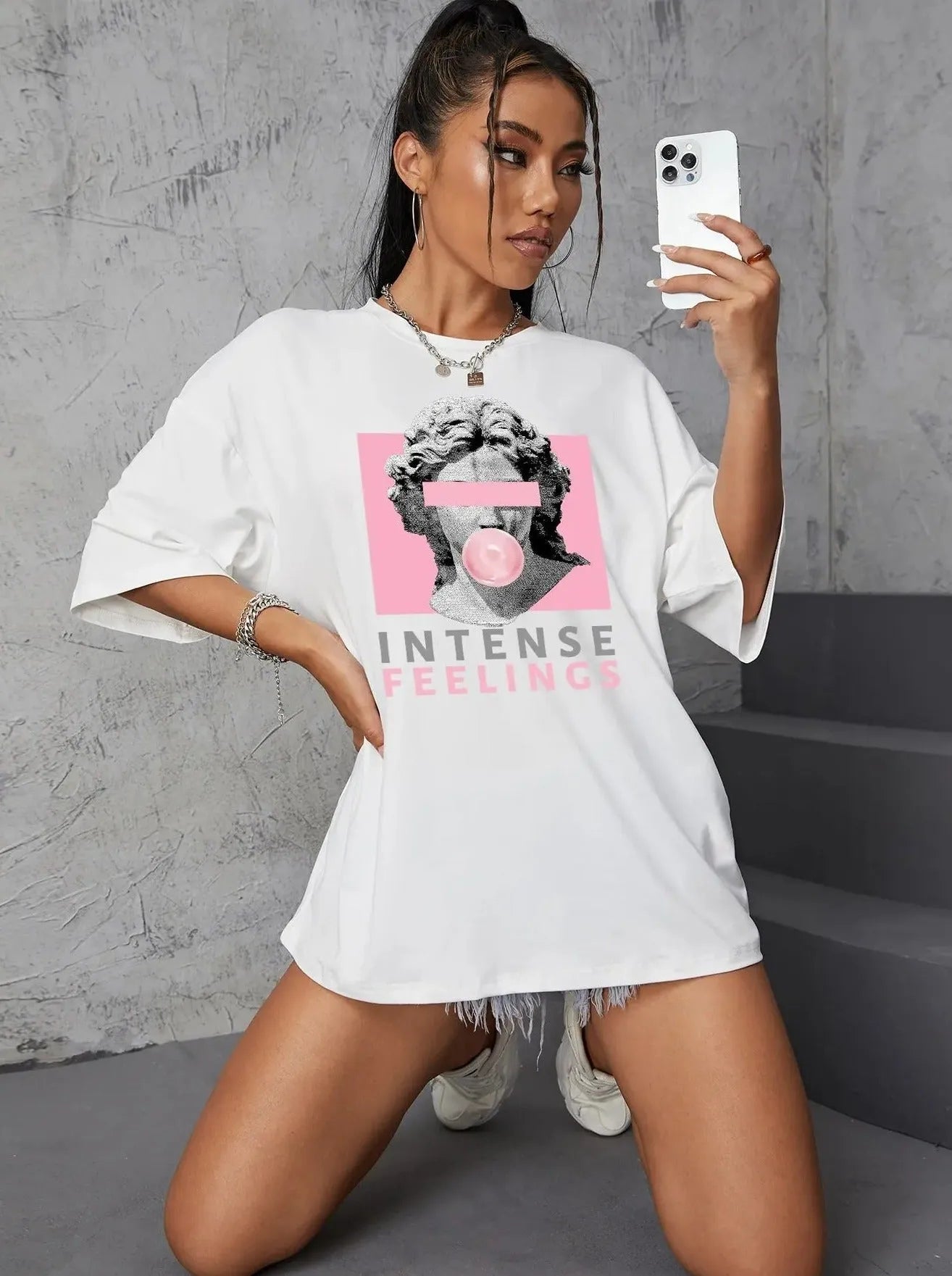 Frau in weißem Oversize-T-Shirt mit "Intense Feelings"-Aufdruck, Smartphone in der Hand, modisch, urbaner Stil, Selfie, Streetwear-Trend.