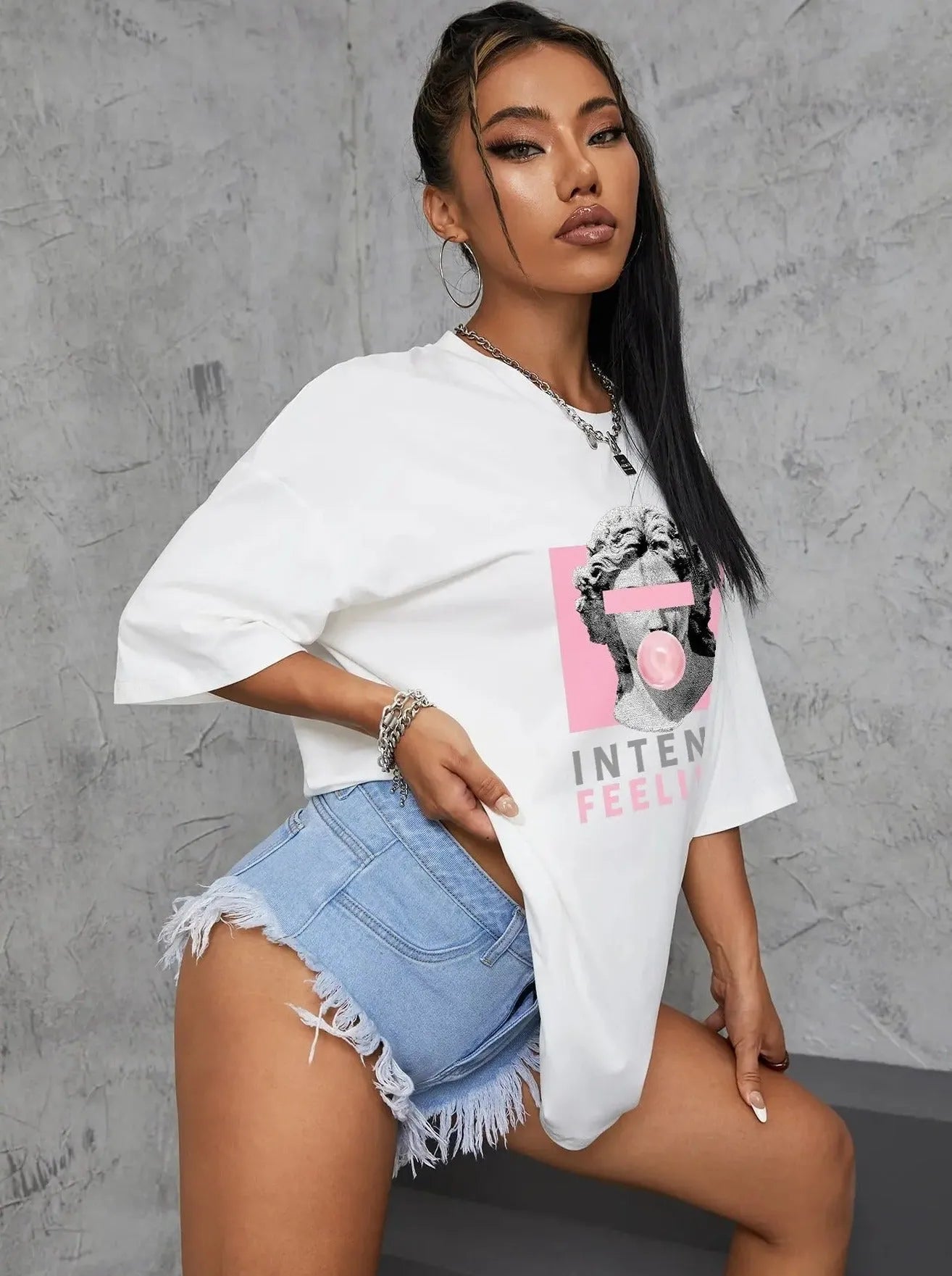 Frau in weißem T-Shirt mit Aufdruck und blauen Jeans-Shorts, posiert vor grauem Hintergrund. Mode, Streetwear, lässiger Look, trendige Kleidung.