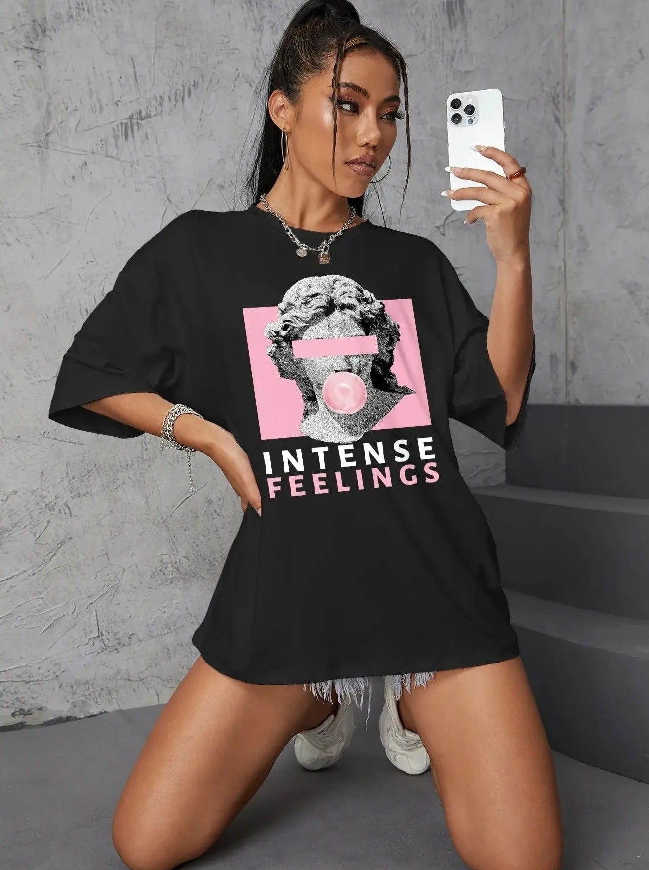 Frau in schwarzem Oversize-T-Shirt mit antikem Skulptur-Print und "Intense Feelings"-Schriftzug, posiert mit Smartphone. Trendige Streetwear-Mode.