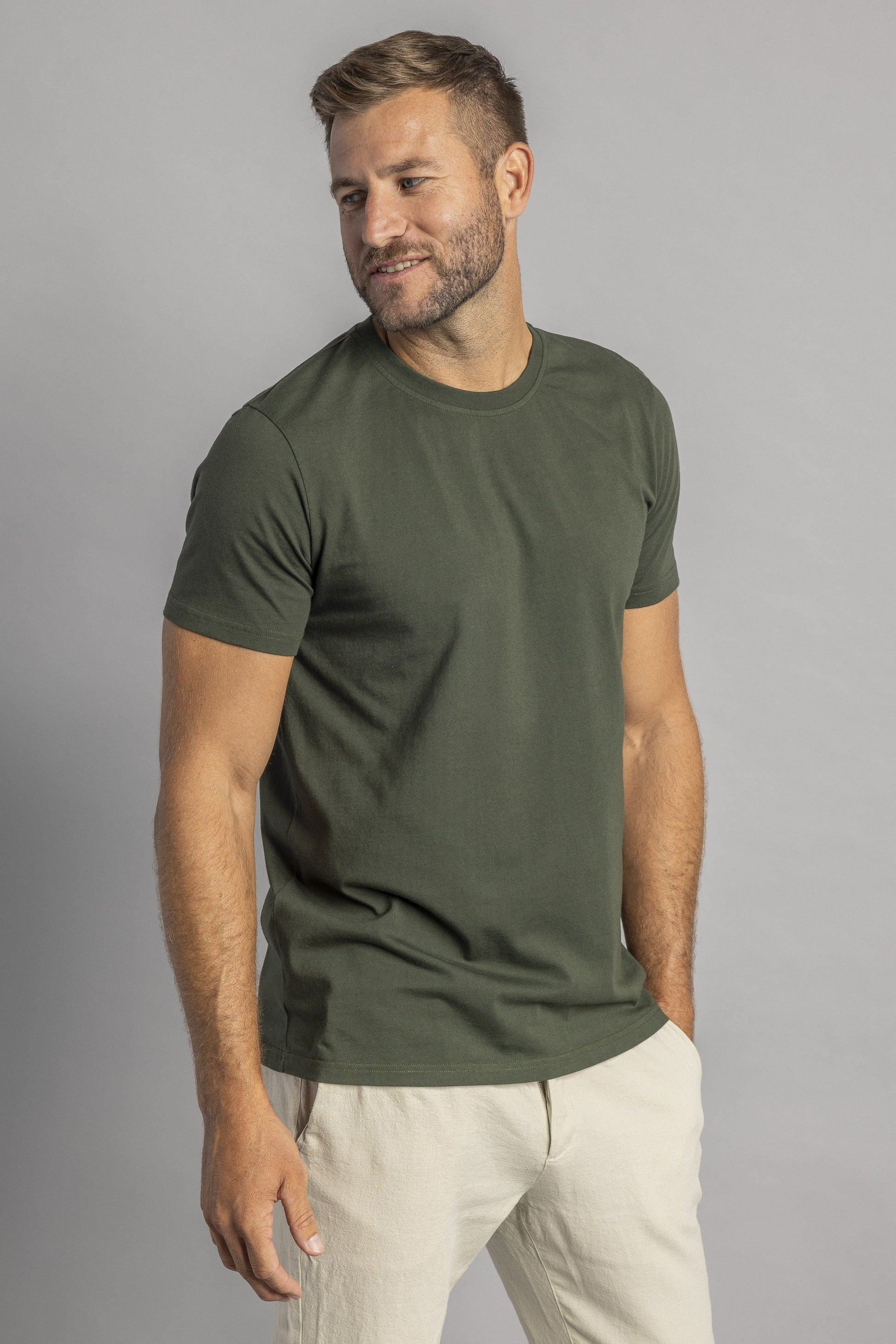 Mann in olivgrünem T-Shirt und beigen Hosen, lächelnd, vor grauem Hintergrund.