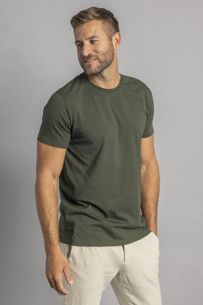 Mann in olivgrünem T-Shirt und beigen Hosen, lächelnd, vor grauem Hintergrund.