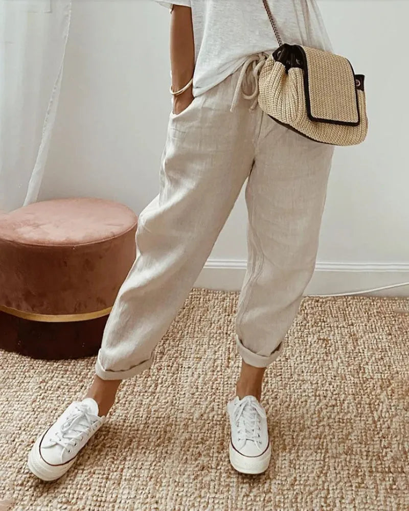 Damenmode: Beige Leinenhose, lässig geschnitten, kombiniert mit weißen Sneakers und einer stilvollen Strohtasche. Perfekt für den Sommerlook.