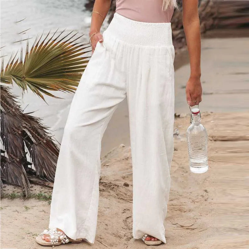 Frau in weißen Leinenhosen am Strand, hält eine Wasserflasche. Sommermode, Strandkleidung, lässiger Look, bequeme Damenhosen.
