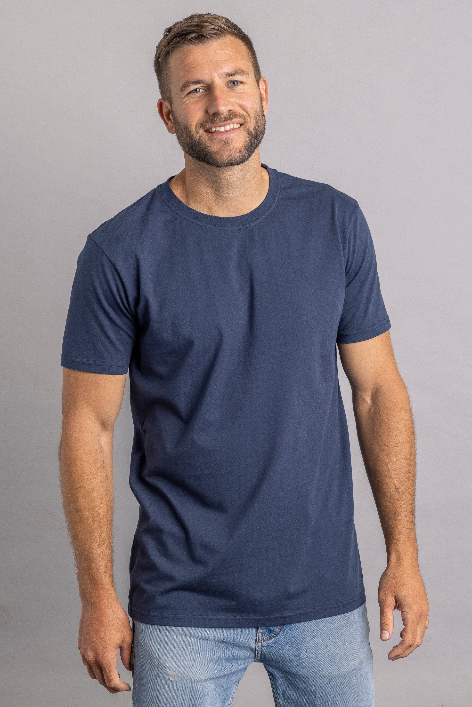 Mann in dunkelblauem T-Shirt und Jeans, lächelnd, vor grauem Hintergrund.