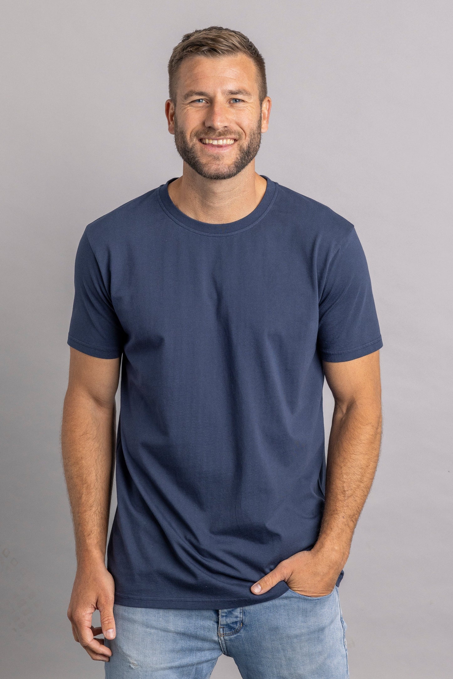 Mann in dunkelblauem T-Shirt und Jeans, lächelnd, vor grauem Hintergrund.