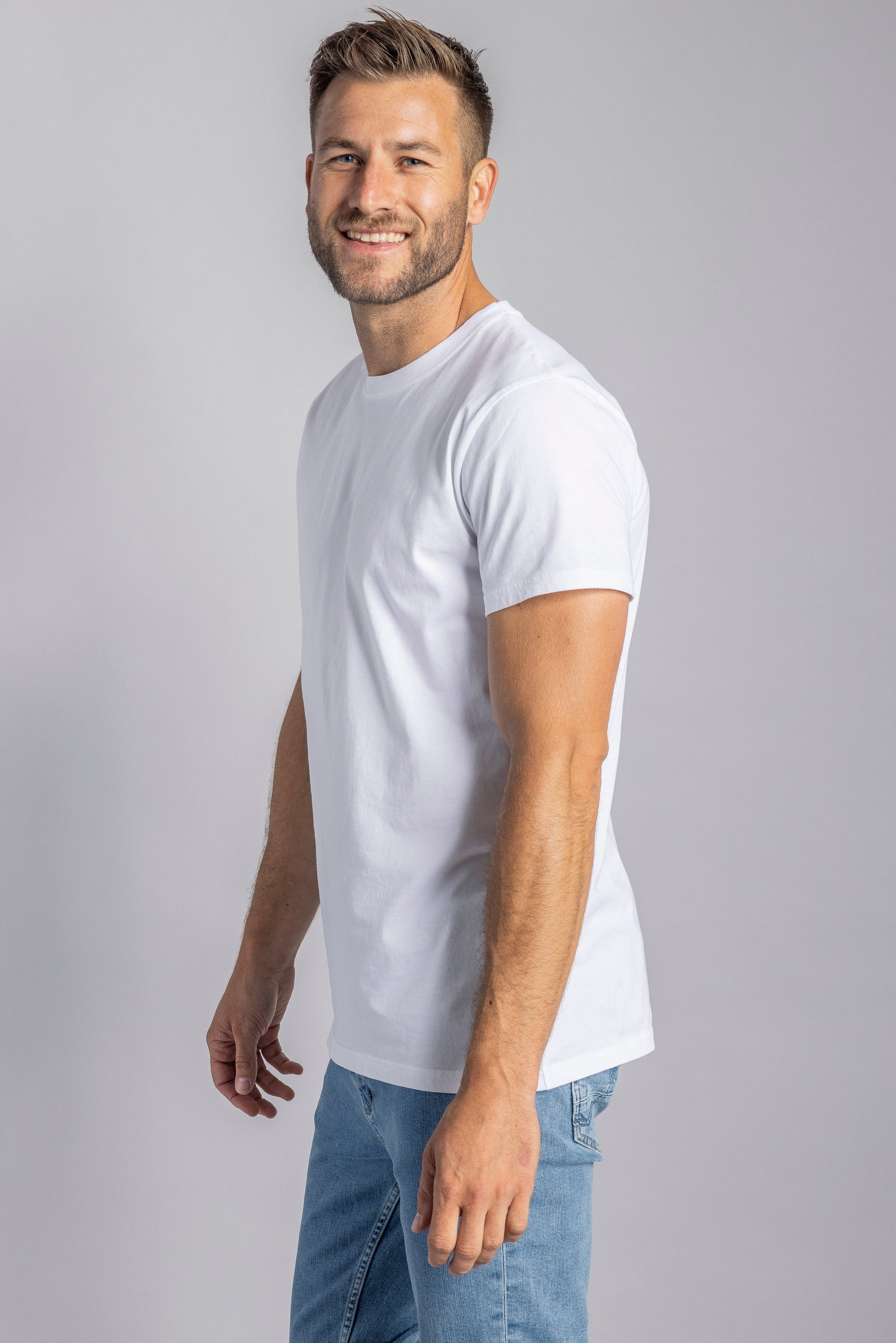 Mann in weißem T-Shirt und blauer Jeans, lächelnd, seitlich stehend, grauer Hintergrund.