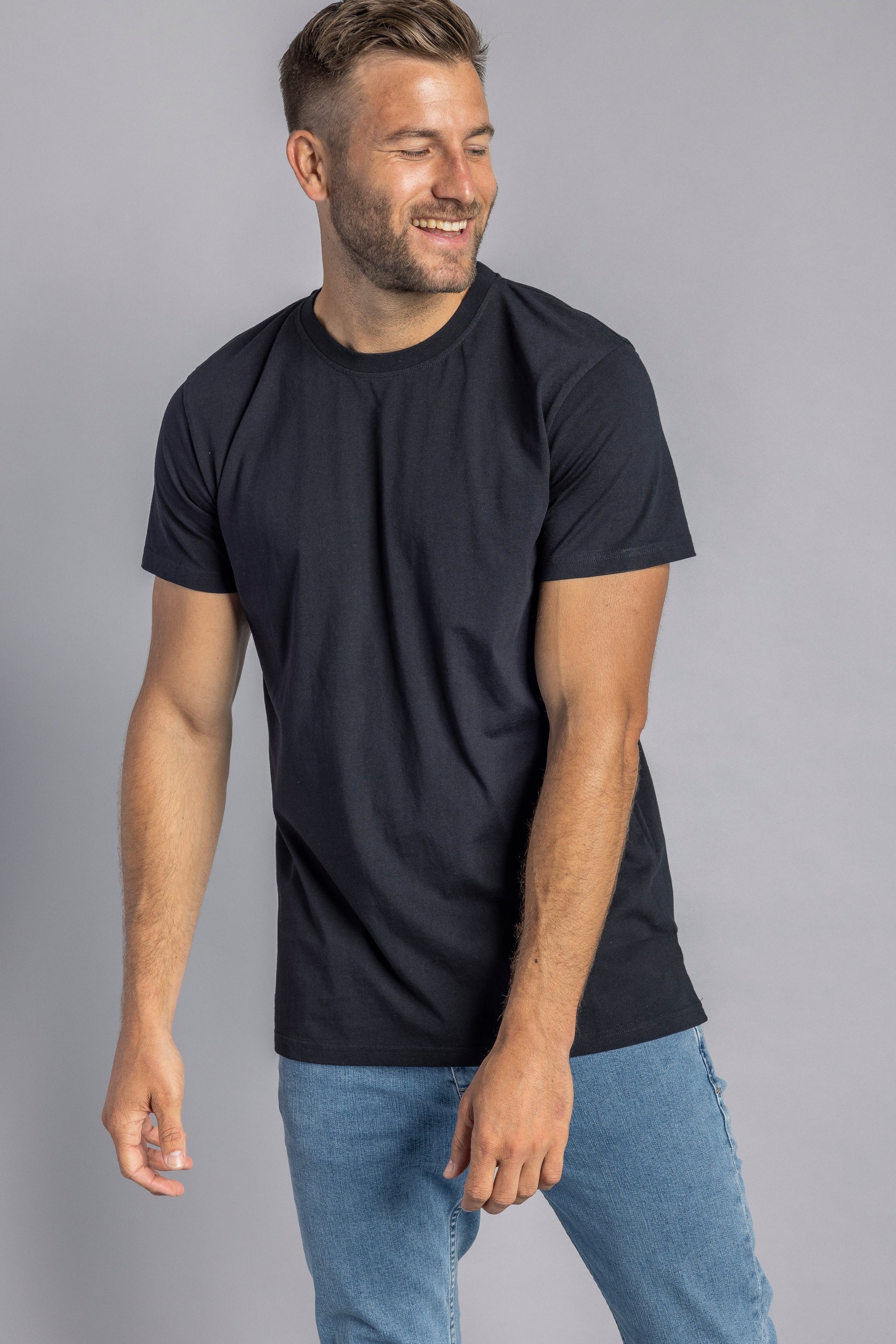Mann in schwarzem T-Shirt und blauer Jeans, lächelnd, vor grauem Hintergrund.