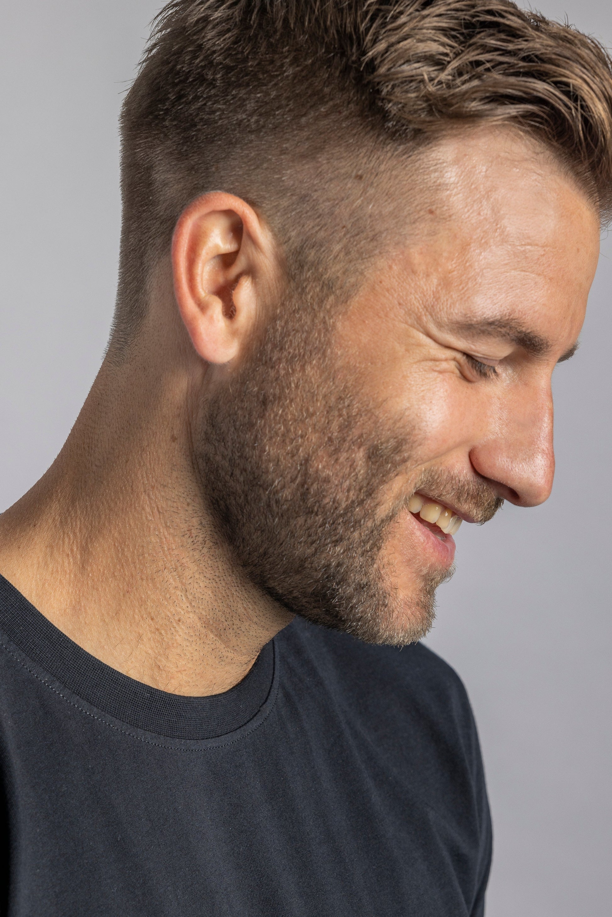 Lächelnder Mann mit braunem Haar in schwarzem T-Shirt, Seitenprofil, grauer Hintergrund.