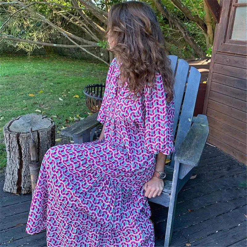 Frau im Boho-Maxi-Kleid mit Laternenärmeln und V-Ausschnitt, sitzt im Garten. Sommermode, Urlaubskleid, femininer Druck, entspannter Stil.