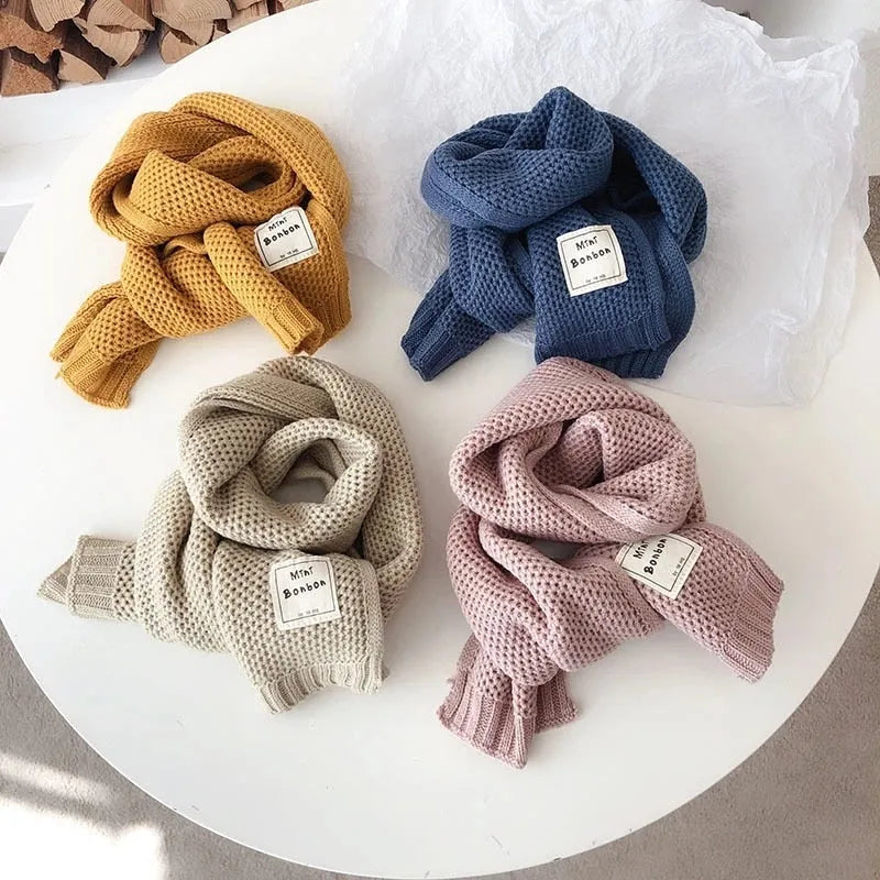 Vier gestrickte Schals in Gelb, Blau, Beige und Rosa auf einem Tisch. Modische Accessoires, warm, weich, ideal für Herbst und Winter.