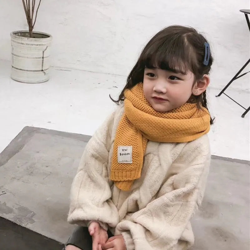 Mädchen mit gelbem Schal und beigem Pullover sitzt auf grauem Boden. Kinderkleidung, Wintermode, gemütlich, stilvoll, modisch.