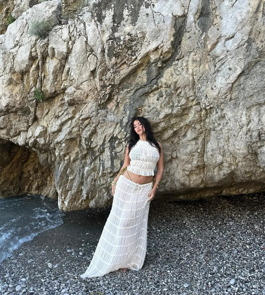 Frau in weißem Sommerkleid posiert am Felsenstrand. Strandmode, Sommeroutfit, Urlaubslook, Naturkulisse, Strandfotografie.