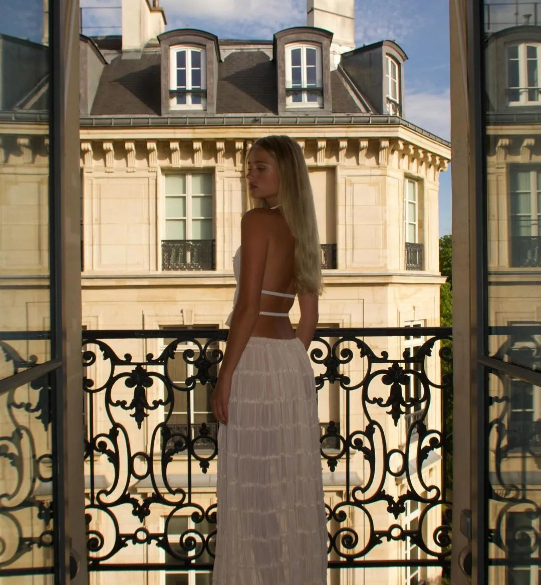 Frau auf Balkon mit weißem Kleid, Blick auf Pariser Architektur. Elegante Mode, Sommerstil, Stadtansicht, französische Eleganz.