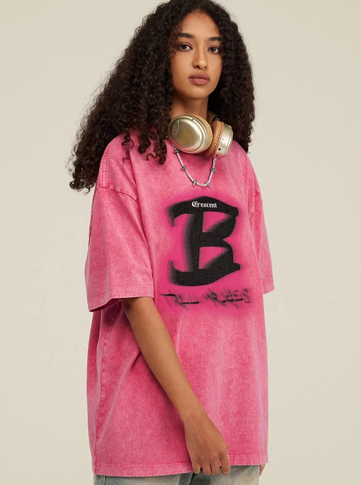 Frau mit lockigem Haar trägt ein pinkes Oversize-T-Shirt mit großem "B"-Logo und Kopfhörer. Modetrend, Streetwear, lässiger Stil.