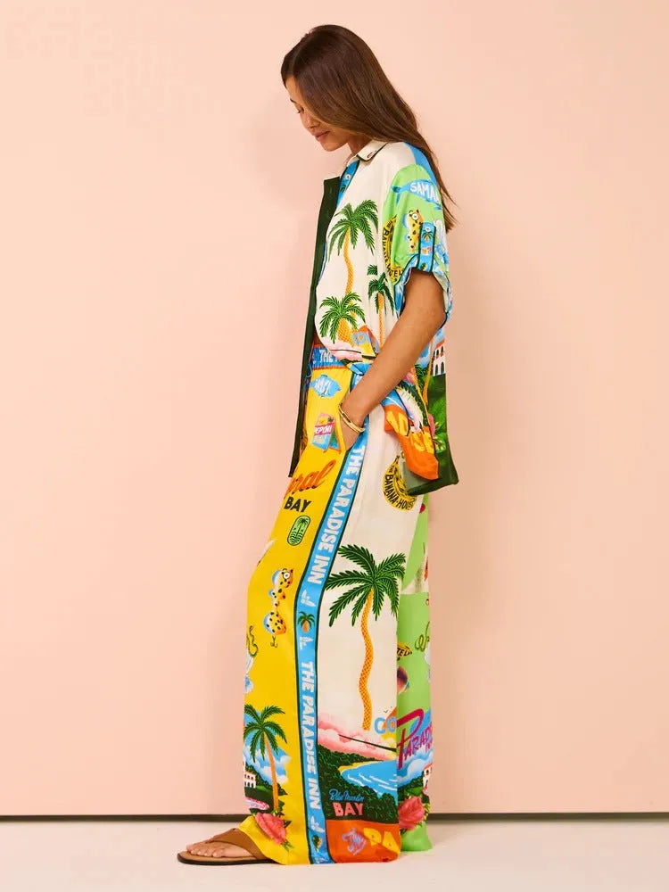 Frau in farbenfrohem, tropischem Maxikleid mit Palmen- und Strandmotiven, ideal für Sommermode und Strandurlaub. Bequeme Sandalen ergänzen den Look.