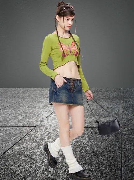 Frau in modischem Outfit: grünes Crop-Top mit Schmetterlingsmotiv, Jeansrock, schwarze Tasche. Trendige Streetwear, jugendlicher Stil.