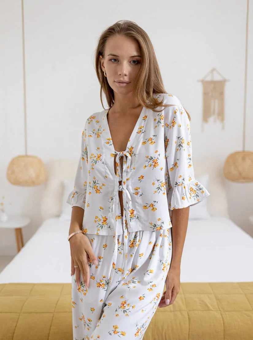 Frau in geblümtem Pyjama-Set, bestehend aus Oberteil mit Rüschenärmeln und passender Hose, steht in modernem Schlafzimmer. Ideal für Nachtwäsche.
