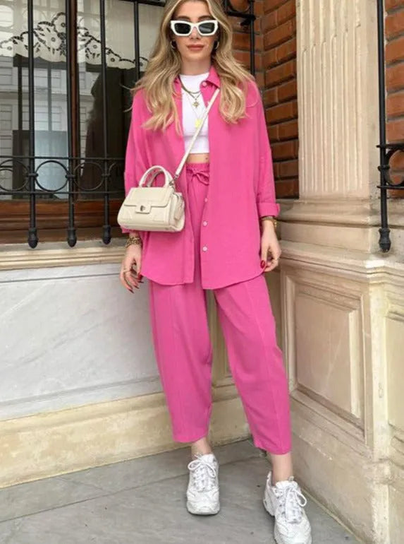 Frau in pinkem Outfit mit weißer Tasche und Sonnenbrille, steht vor Backsteinwand. Modetrend, Streetstyle, lässig, urbaner Look, Sommermode.