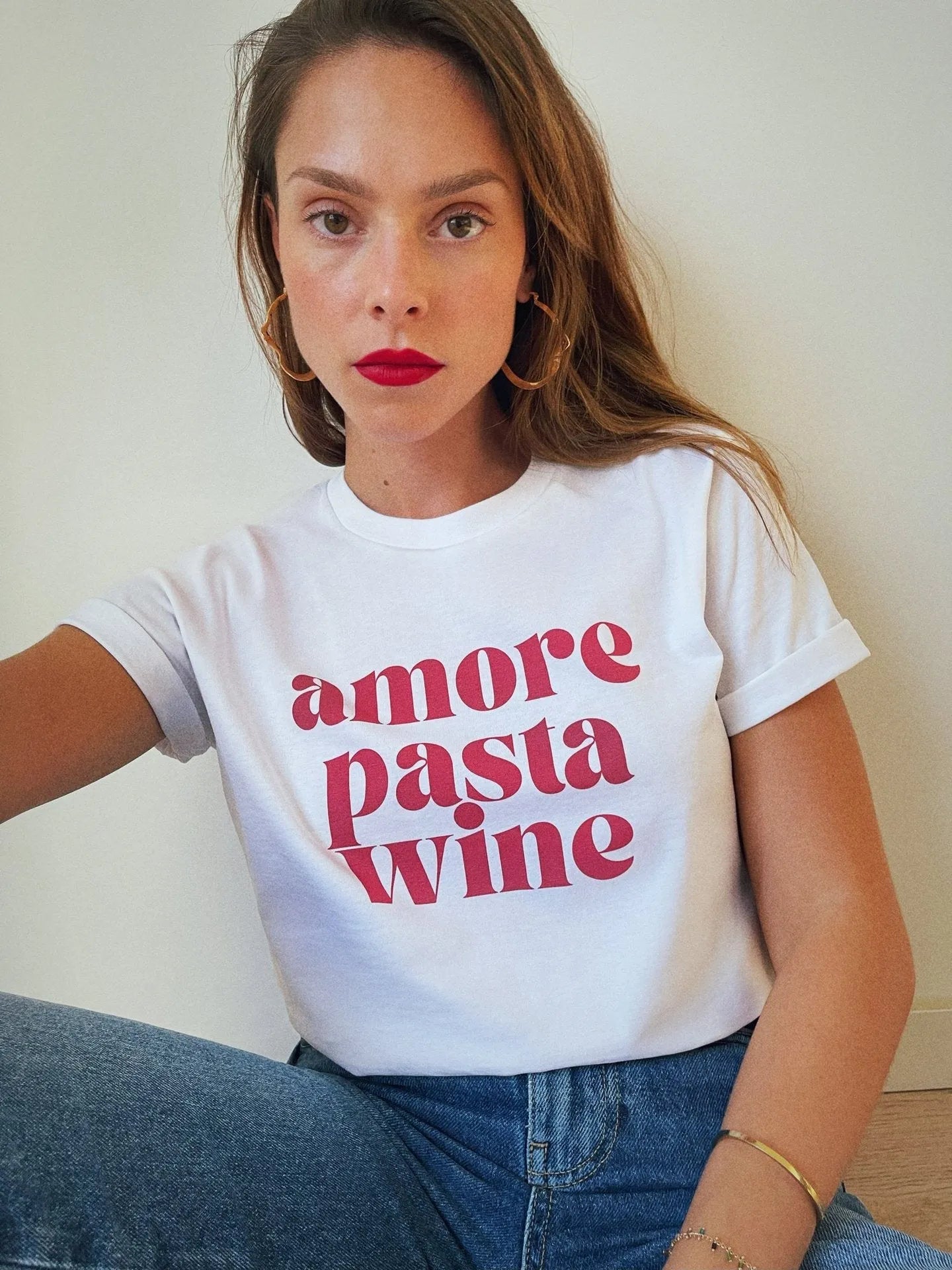 Frau trägt weißes T-Shirt mit rotem Aufdruck "amore pasta wine", kombiniert mit blauen Jeans. Modetrend, lässiger Stil, Statement-Shirt.
