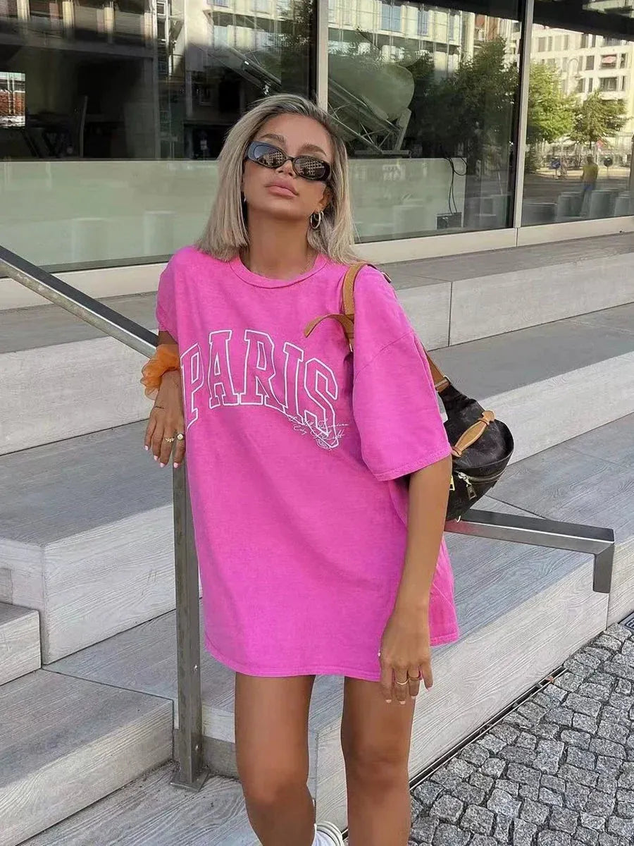 Frau in pinkem "Paris" Oversize-T-Shirt, Sonnenbrille, blonden Haaren, posiert lässig auf Treppe. Modetrend, Streetstyle, Sommerlook.