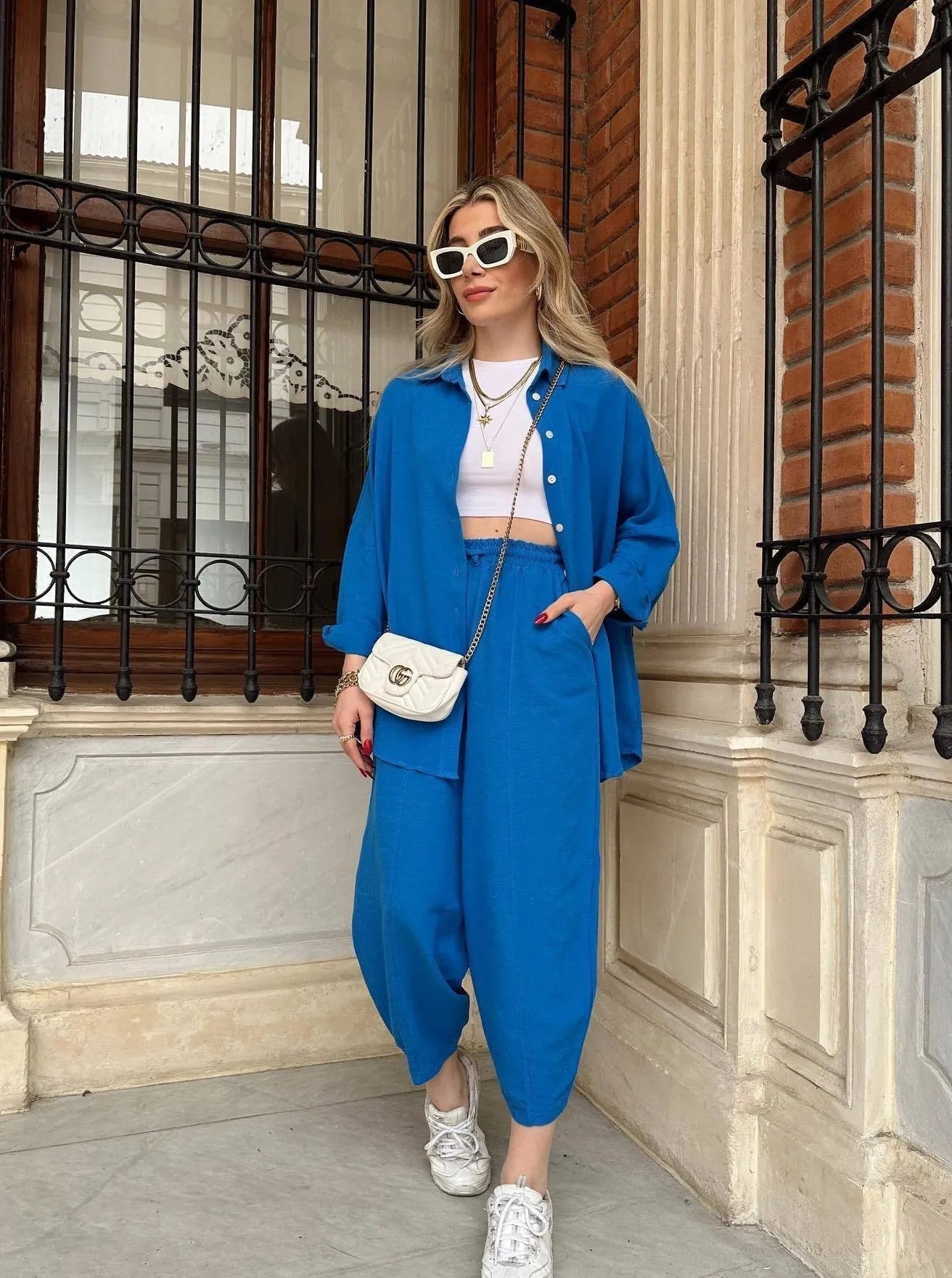 Frau in blauem Oversize-Anzug, weißem Crop-Top und Sonnenbrille, posiert vor Backsteinwand. Modetrend, Streetstyle, lässiger Look.