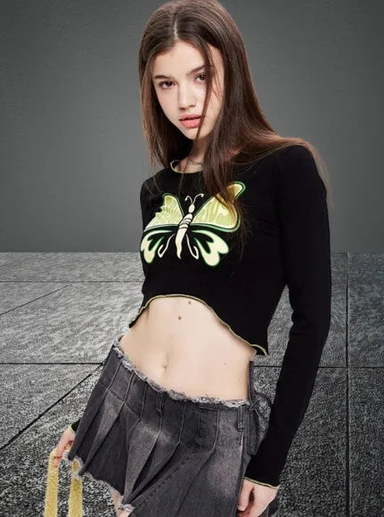 Junge Frau in modischem, schwarzem Crop-Top mit Schmetterlingsmotiv und grauem Faltenrock. Trendige Streetwear für stilbewusste Frauen.