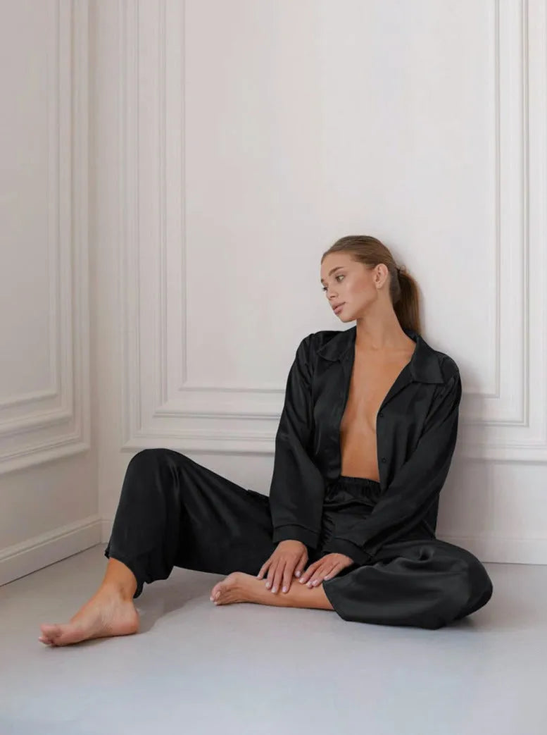 Frau in schwarzem Satin-Pyjama sitzt entspannt auf dem Boden. Elegante Nachtwäsche, luxuriöses Design, gemütliche Loungewear, stilvolle Mode.