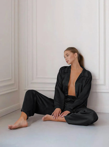 Frau in schwarzem Satin-Pyjama sitzt entspannt auf dem Boden. Elegante Nachtwäsche, luxuriöses Design, gemütliche Loungewear, stilvolle Mode.