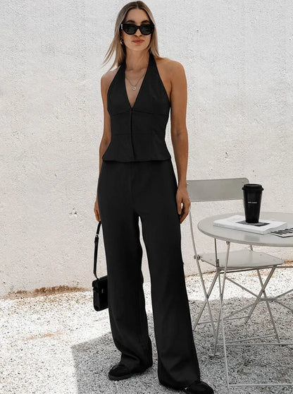 Frau in schwarzem Jumpsuit, Sonnenbrille und Handtasche, steht neben weißem Tisch mit Kaffeebecher. Elegante Mode, Sommeroutfit, Streetstyle.