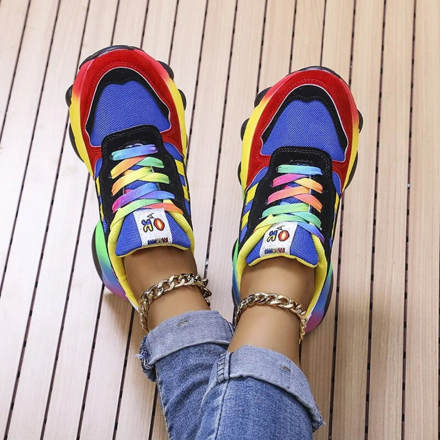 Bunte Sneaker mit Regenbogen-Schnürsenkeln, auffälligem Design und goldenen Knöchelketten auf Holzuntergrund. Modische Schuhe, trendige Accessoires.