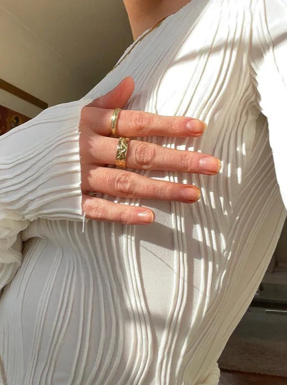 Frau in weißem, plissiertem Oberteil zeigt goldene Ringe. Elegante Mode, minimalistisch, Schmuck, Stil, Trend, Damenbekleidung, Accessoires.