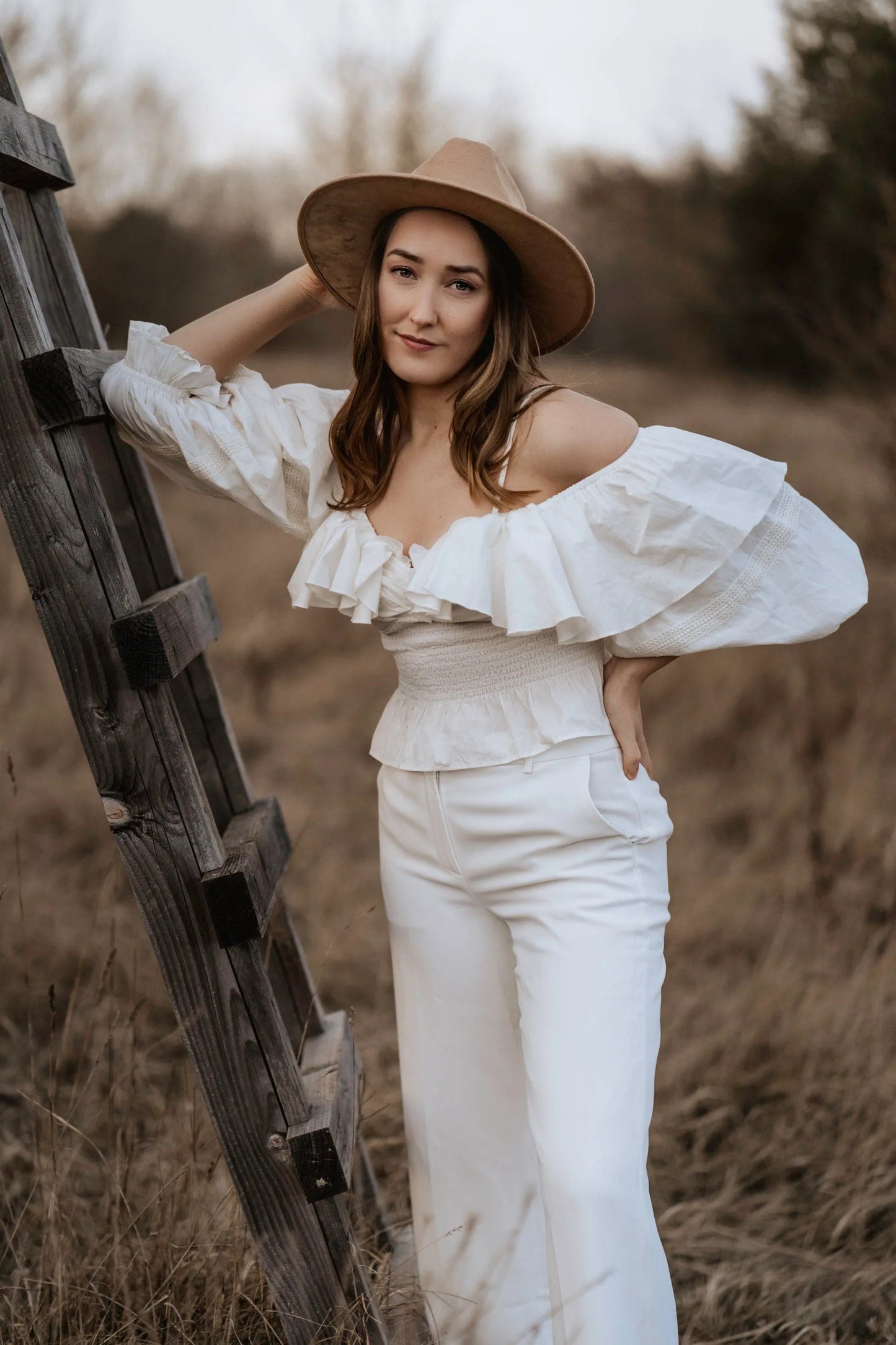 Frau in weißem, schulterfreiem Kleid mit Rüschen und Hut lehnt an Holzleiter im Freien. Mode, Boho-Stil, Outdoor-Fotografie, Herbst.