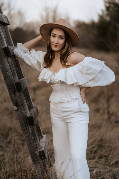 Frau in weißem, schulterfreiem Kleid mit Rüschen und Hut lehnt an Holzleiter im Freien. Mode, Boho-Stil, Outdoor-Fotografie, Herbst.