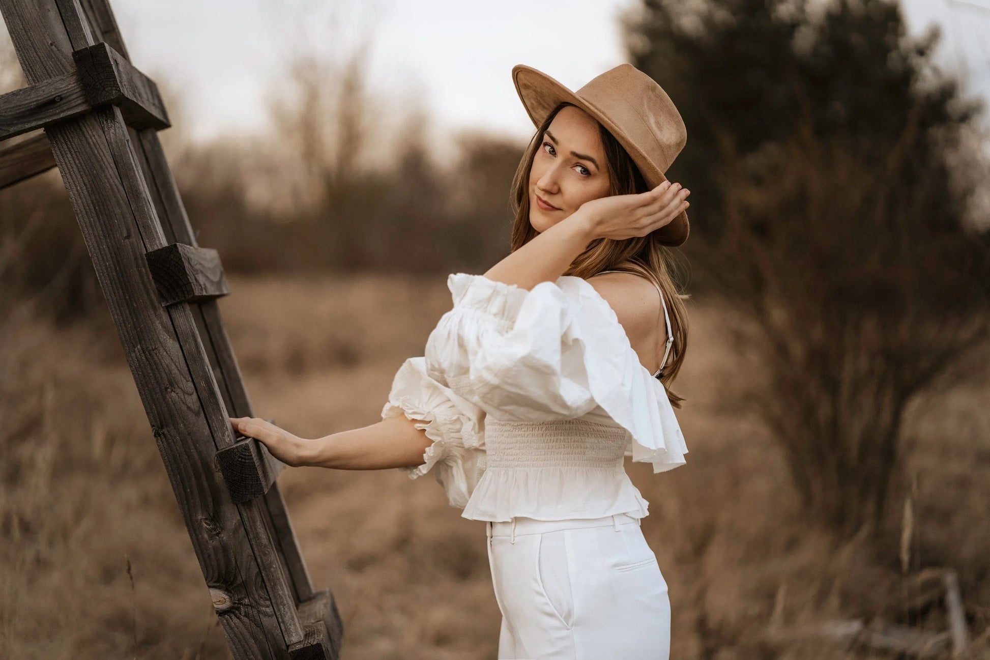 Junge Frau in weißer Off-Shoulder-Bluse und Hut lehnt an Holzleiter im Freien. Modetrend, Boho-Stil, Natur, Herbstmode, Damenbekleidung.