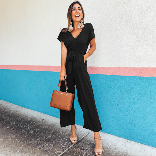Frau im schwarzen Jumpsuit mit brauner Handtasche, lächelnd vor bunter Wand.