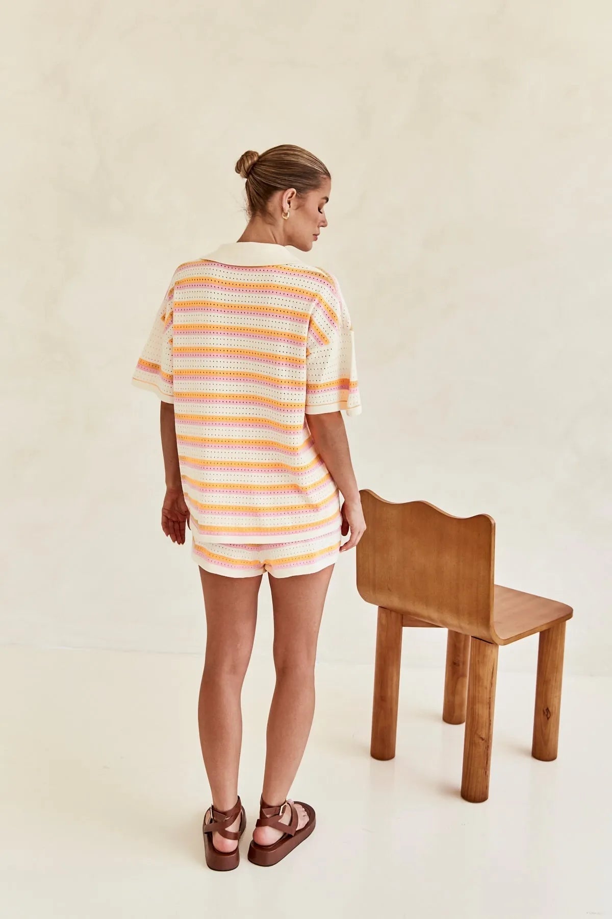 Frau in gestreiftem, pastellfarbenem Strick-Set mit Shorts und T-Shirt, steht neben einem Holzstuhl. Sommermode, lässiger Look, trendige Damenbekleidung.